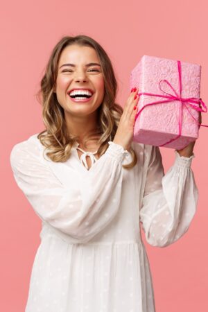 Holidays, celebration and women concept. Portrait of happy charismatic blond girl shaking gift box wondering whats inside as celebrating birthday, receive b-day presents, pink background