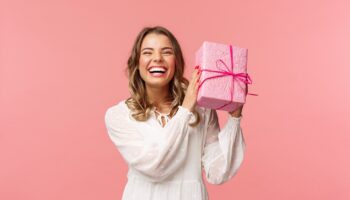 Holidays, celebration and women concept. Portrait of happy charismatic blond girl shaking gift box wondering whats inside as celebrating birthday, receive b-day presents, pink background