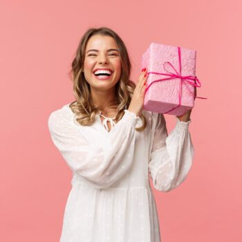 Holidays, celebration and women concept. Portrait of happy charismatic blond girl shaking gift box wondering whats inside as celebrating birthday, receive b-day presents, pink background